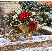 Load image into Gallery viewer, A small wooden wheelbarrow filled with a festive Christmas arrangement, including red poinsettias, white flowers, holly, pinecones, and red berries, set outdoors on a snow-dusted stone surface.
